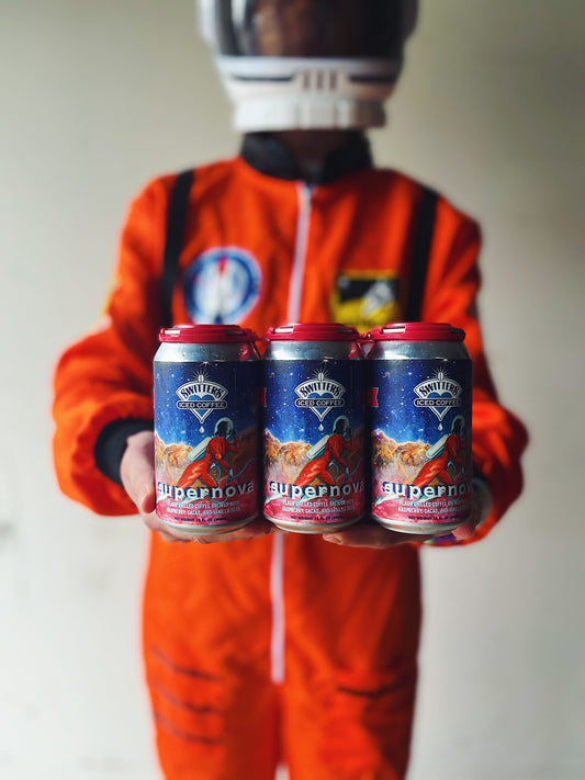 Sheri the astronaut looks off into the cosmic distance as she holds a 6 pack of Switters Supernova iced coffee in one hand, and her helmet in the other. Behind her is a backdrop of abstract space